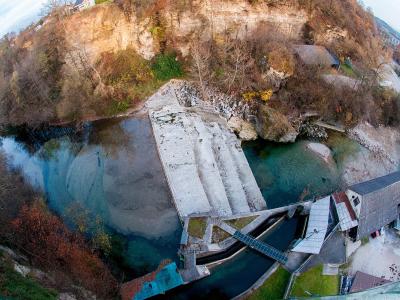 An aerial fisheye view of a river gorge featuring a concrete weir or dam structure, a small building with a grey roof, and steep rocky cliffs with autumn foliage.