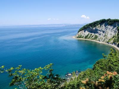 A scenic coastal view showing a high, steep cliff with layered rock formations and green vegetation on top, curving down to a sandy beach and turquoise water under a clear blue sky.