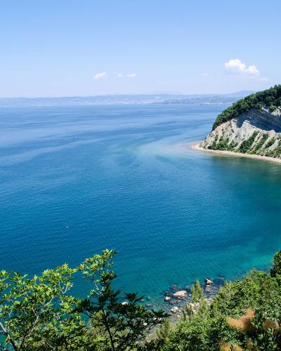 A scenic coastal view showing a high, steep cliff with layered rock formations and green vegetation on top, curving down to a sandy beach and turquoise water under a clear blue sky.
