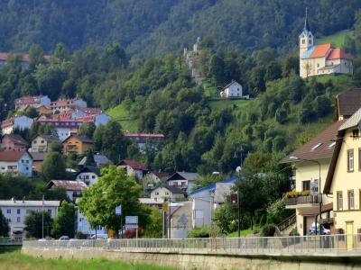 A view of a town built on a steep, forested hillside, featuring a prominent church with a red roof and a line of chapels (a calvary) leading up the slope, with a river embankment and road in the foreground.