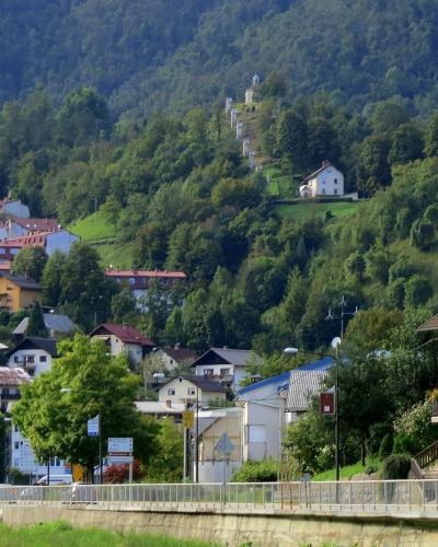 A view of a town built on a steep, forested hillside, featuring a prominent church with a red roof and a line of chapels (a calvary) leading up the slope, with a river embankment and road in the foreground.