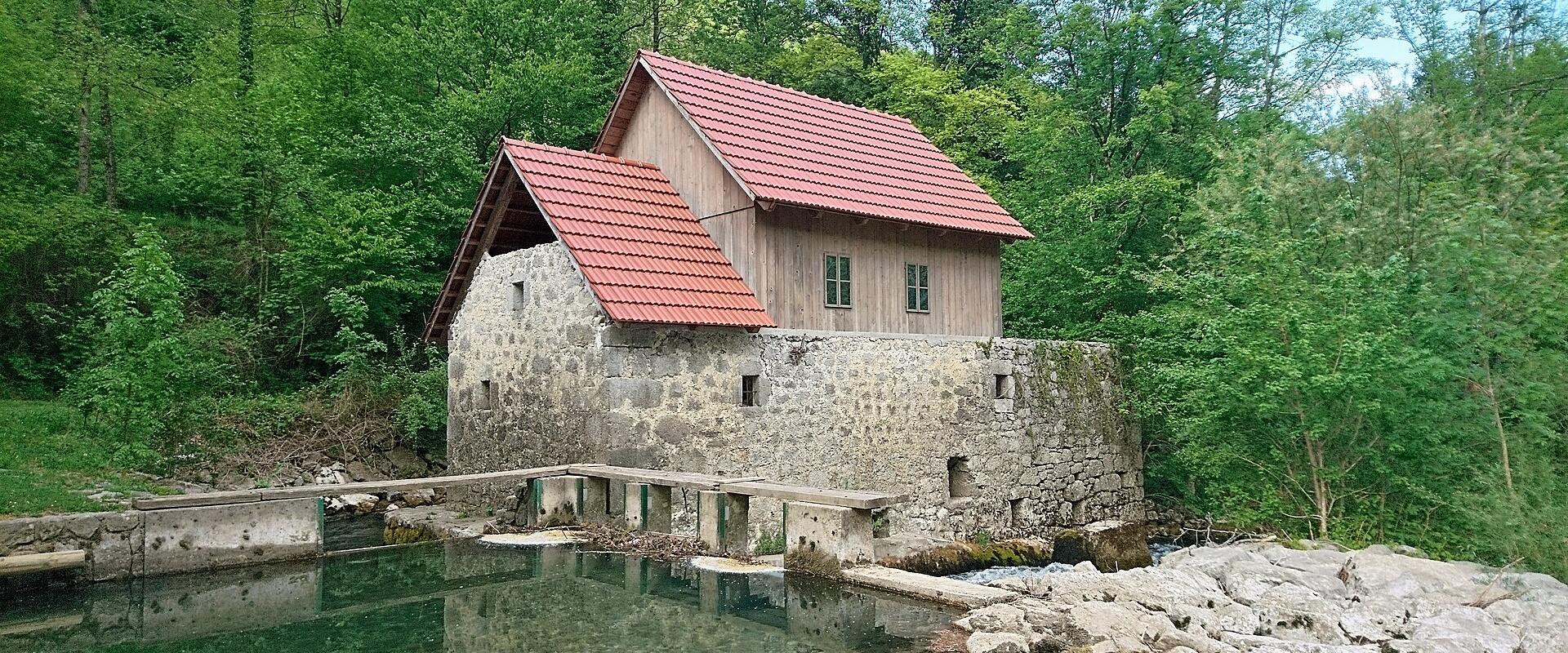 A stone and wood building with a red tiled roof situated on the bank of a clear, green river, featuring a concrete weir or dam structure in the foreground.