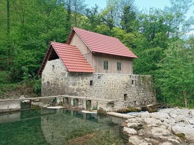 A stone and wood building with a red tiled roof situated on the bank of a clear, green river, featuring a concrete weir or dam structure in the foreground.