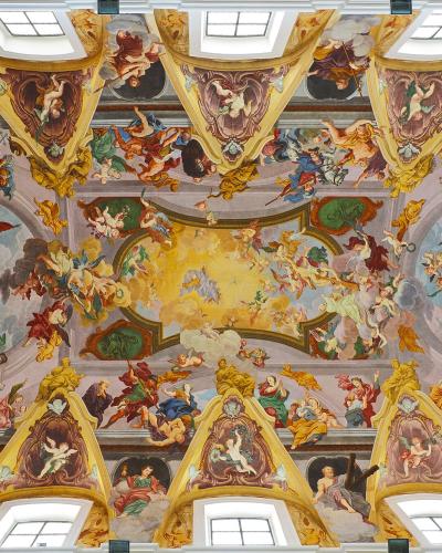 The image shows an ornate Baroque ceiling fresco with numerous figures, cherubs, and architectural illusions, featuring skylights and gilded stucco work.