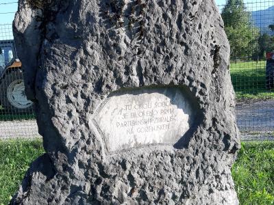 A large, rough-hewn stone monument with an engraved plaque reading "TU OKOLI ŠOBCE JE BILO ENO PRVIH PARTIZANSKIH ZBIRALIŠČ NA GORENJSKEM" (Here around Šobec was one of the first partisan gathering places in Gorenjska), situated outdoors near a fence