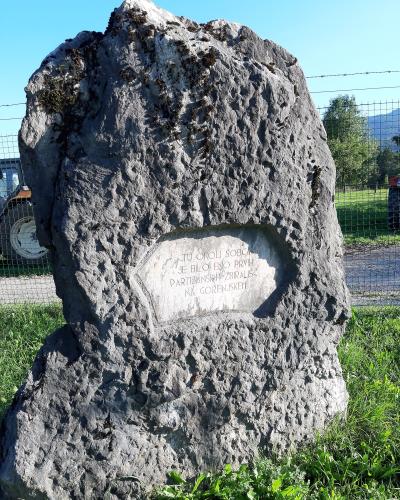 A large, rough-hewn stone monument with an engraved plaque reading "TU OKOLI ŠOBCE JE BILO ENO PRVIH PARTIZANSKIH ZBIRALIŠČ NA GORENJSKEM" (Here around Šobec was one of the first partisan gathering places in Gorenjska), situated outdoors near a fence