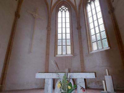 The interior of a small, Gothic-style chapel featuring a stone altar, a wooden cross on the wall, tall arched windows, and a terracotta tiled floor.