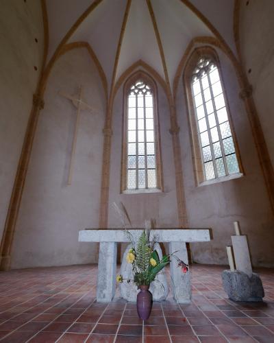 The interior of a small, Gothic-style chapel featuring a stone altar, a wooden cross on the wall, tall arched windows, and a terracotta tiled floor.