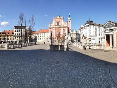 A wide-angle view of a paved public square featuring a prominent pink Baroque church with a bell tower, flanked by classical buildings and a stone balustrade, under a blue sky.
