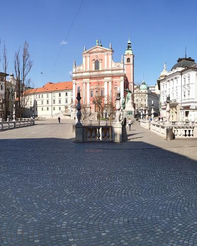 A wide-angle view of a paved public square featuring a prominent pink Baroque church with a bell tower, flanked by classical buildings and a stone balustrade, under a blue sky.