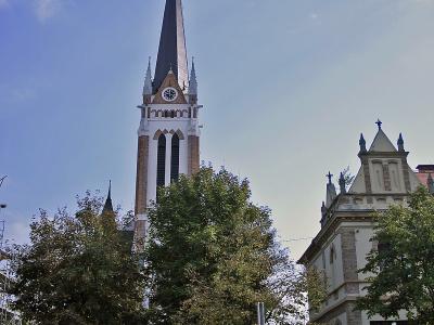 A tall, white and brick neo-Gothic church tower with a dark spire, clock face, and cross, viewed from a street with trees, a fence, and a parked car.