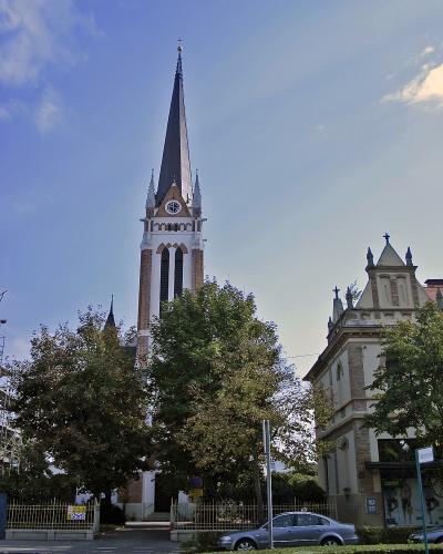A tall, white and brick neo-Gothic church tower with a dark spire, clock face, and cross, viewed from a street with trees, a fence, and a parked car.