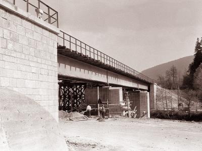 A black and white photograph showing the construction of a railway bridge with stone masonry abutments and steel girder spans, supported by temporary wooden trestling, with workers visible on the ground.