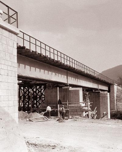 A black and white photograph showing the construction of a railway bridge with stone masonry abutments and steel girder spans, supported by temporary wooden trestling, with workers visible on the ground.