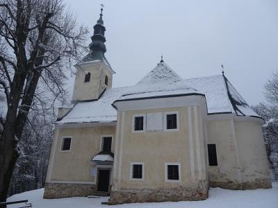 A yellow church with a dark spire and snow-covered roof, surrounded by bare trees in a snowy landscape.