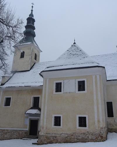 A yellow church with a dark spire and snow-covered roof, surrounded by bare trees in a snowy landscape.