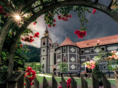 A view through a wooden archway of red roses looking towards a large monastery complex featuring a white church tower and a grey building with white quatrefoil patterns, set against a dramatic sky.