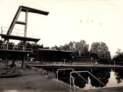 A black-and-white photograph showing an outdoor swimming pool complex with a tall concrete diving tower, lane markers, and a long low building in the background surrounded by trees.