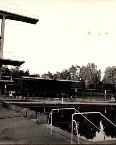 A black-and-white photograph showing an outdoor swimming pool complex with a tall concrete diving tower, lane markers, and a long low building in the background surrounded by trees.