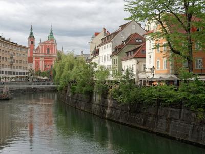 The image shows a view of the Ljubljanica River in Ljubljana, Slovenia, with the distinctive pink facade and twin green spires of the Franciscan Church of the Annunciation visible in the background on the left bank.