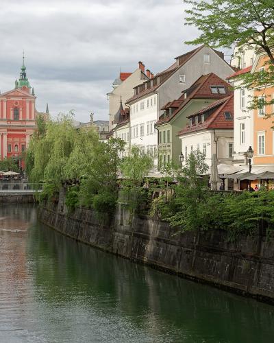 The image shows a view of the Ljubljanica River in Ljubljana, Slovenia, with the distinctive pink facade and twin green spires of the Franciscan Church of the Annunciation visible in the background on the left bank.