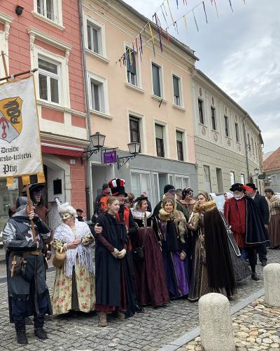 A group of people in historical costumes and masks standing on a cobblestone street in front of colorful buildings, with one person holding a banner that reads "Društvo Cesarsko-králevski Ptuj".