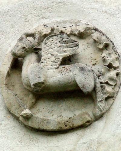 A close-up photograph of a weathered stone relief carving depicting a winged bull (or griffin-like creature) set within a circular frame on a rough, light-colored wall.