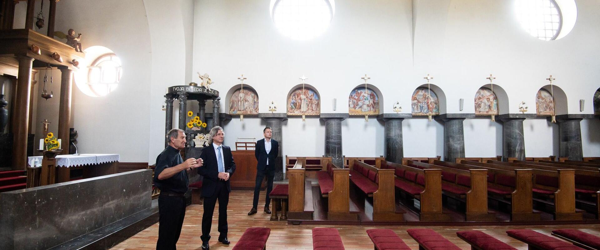 Two men, one in a suit and one in clerical clothing, standing indoors next to a large marble column and wooden furniture, looking at a book.