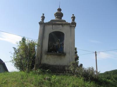 A Baroque-style roadside chapel or wayside shrine situated on a grassy slope, featuring a painted religious scene within an arched niche and topped with decorative urns.