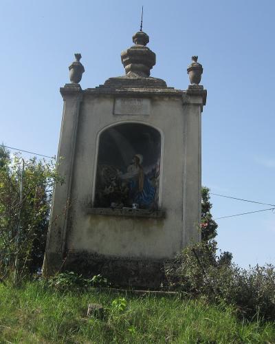 A Baroque-style roadside chapel or wayside shrine situated on a grassy slope, featuring a painted religious scene within an arched niche and topped with decorative urns.