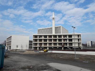 A modern white building complex with a tall minaret and a distinctive geometric lattice screen on the upper level, captured during construction or landscaping with piles of dirt and a yellow vehicle in the foreground.