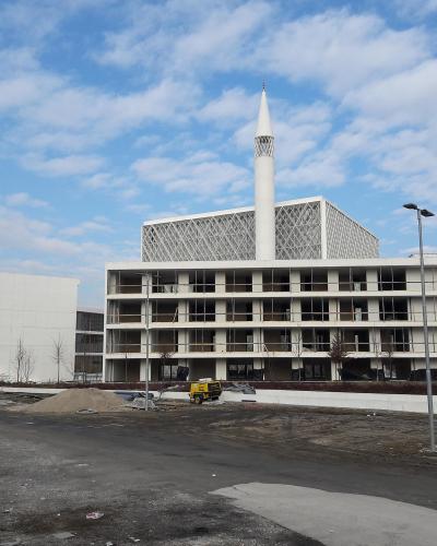 A modern white building complex with a tall minaret and a distinctive geometric lattice screen on the upper level, captured during construction or landscaping with piles of dirt and a yellow vehicle in the foreground.