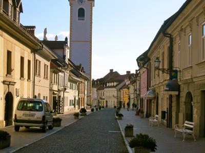 A cobblestone street lined with pastel-colored historic buildings and white benches, leading toward a tall, pale church tower with a dark baroque-style spire and clock face under a clear blue sky.