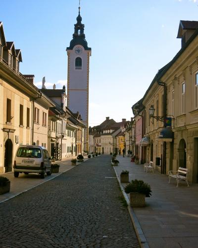 A cobblestone street lined with pastel-colored historic buildings and white benches, leading toward a tall, pale church tower with a dark baroque-style spire and clock face under a clear blue sky.