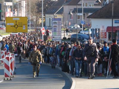 A large crowd of migrants and refugees walking along a road, escorted by Slovenian military and police personnel, with a signpost indicating the border crossing into Slovenia.