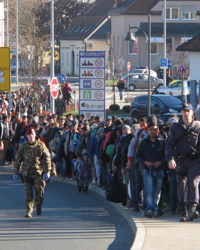A large crowd of migrants and refugees walking along a road, escorted by Slovenian military and police personnel, with a signpost indicating the border crossing into Slovenia.