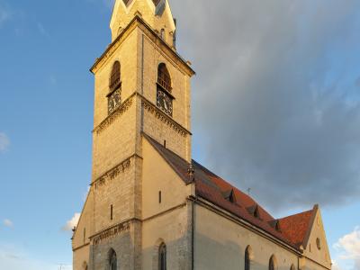 A tall, light-colored stone church with a prominent square tower topped by a dark spire and weather vane, situated in a paved town square with parked cars and outdoor seating.