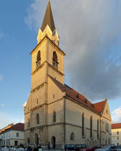 A tall, light-colored stone church with a prominent square tower topped by a dark spire and weather vane, situated in a paved town square with parked cars and outdoor seating.