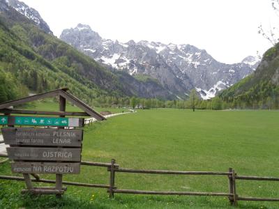 A wide green alpine meadow in a U-shaped valley with snow-capped mountains in the background, featuring a wooden directional signpost in the foreground listing local tourist farms and a pension.