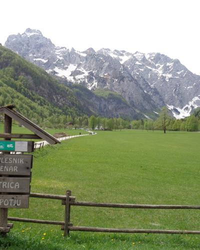 A wide green alpine meadow in a U-shaped valley with snow-capped mountains in the background, featuring a wooden directional signpost in the foreground listing local tourist farms and a pension.