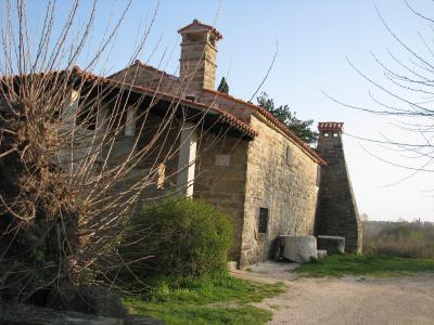 A rustic stone building with a terracotta-tiled roof, two prominent stone chimneys, and a gravel path in the foreground, partially obscured by bare branches in the immediate left foreground.