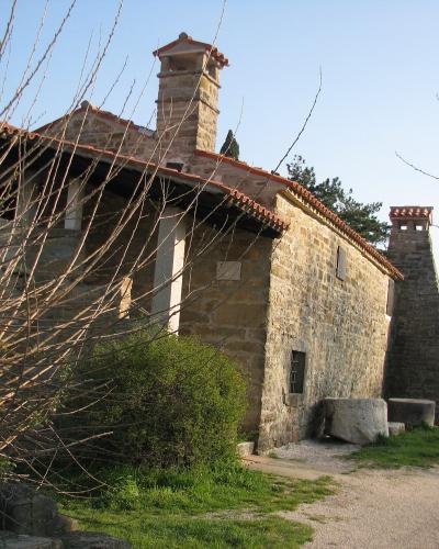 A rustic stone building with a terracotta-tiled roof, two prominent stone chimneys, and a gravel path in the foreground, partially obscured by bare branches in the immediate left foreground.