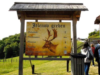 A wooden entrance sign for "Jelenov greben" featuring a stag illustration and multilingual welcome text, set in a grassy, rural landscape with visitors nearby.