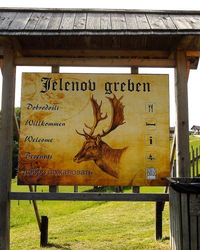 A wooden entrance sign for "Jelenov greben" featuring a stag illustration and multilingual welcome text, set in a grassy, rural landscape with visitors nearby.