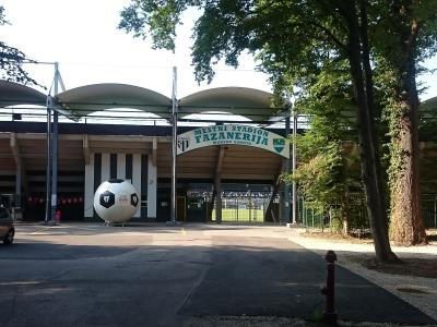The image shows the entrance to a sports stadium with a large soccer ball sculpture, covered stands, and a sign reading "MESTNI STADION FAZANERIJA MURSKA SOBOTA".
