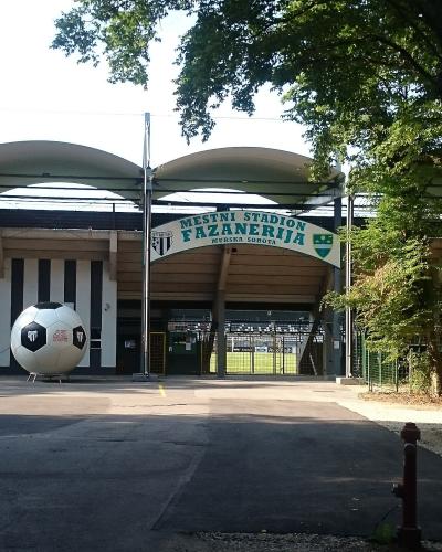 The image shows the entrance to a sports stadium with a large soccer ball sculpture, covered stands, and a sign reading "MESTNI STADION FAZANERIJA MURSKA SOBOTA".