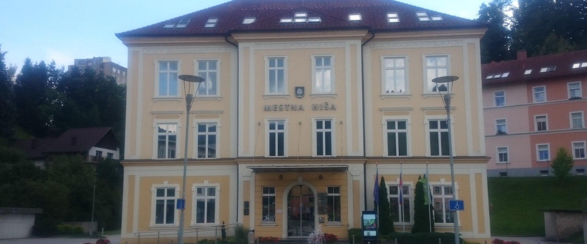A yellow, three-story municipal building with a red tiled roof, identified by the text "MESTNA HIŠA" (Town Hall) above the entrance, situated in a paved town square with street lamps and a small fountain.