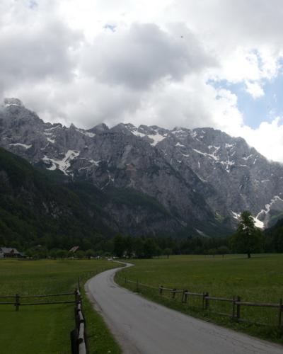 A winding paved road leads through a lush green alpine meadow flanked by wooden fences, with traditional houses on the left and steep, rocky mountain peaks rising in the background under a cloudy sky.