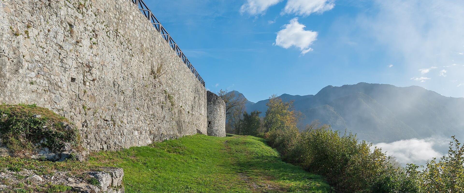 A stone fortress wall with a modern wooden railing runs along a grassy slope, overlooking a valley with mountains and low-lying clouds in the background.