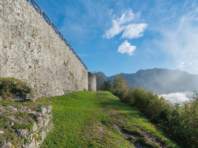 A stone fortress wall with a modern wooden railing runs along a grassy slope, overlooking a valley with mountains and low-lying clouds in the background.